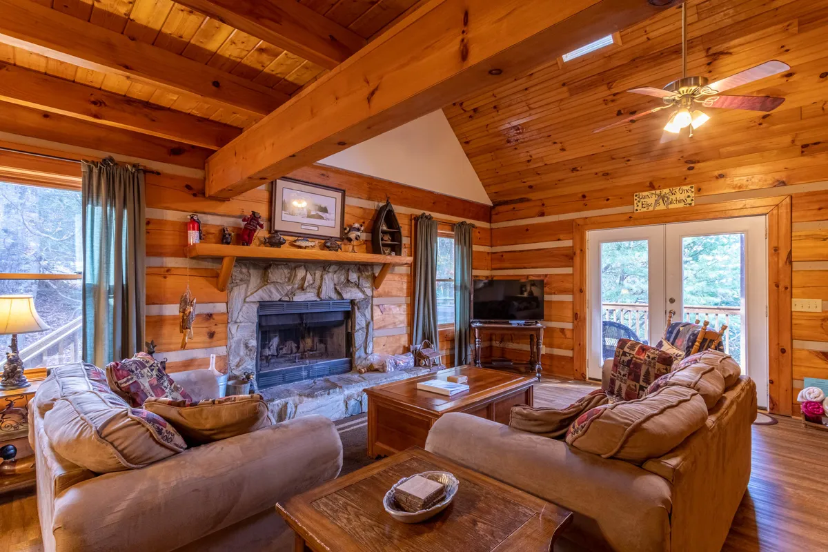 A view of the inviting cabin living area, featuring a cozy fireplace flanked by large windows that let in natural light and connect the space to the outdoors. Plush sofas and rustic wooden beams create a warm, communal atmosphere ideal for relaxing together after a day of mountain adventures.