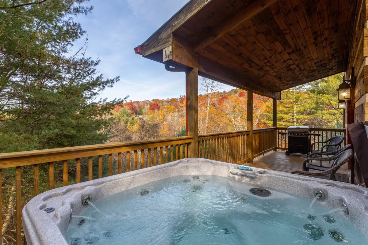 The hero image showcases the exterior of a classic log cabin surrounded by vibrant autumn foliage, with a steaming hot tub set on a wooden deck overlooking rolling hills and the tranquil New River valley. The inviting space hints at cozy evenings soaking under the stars and peaceful mornings spent with nature all around.