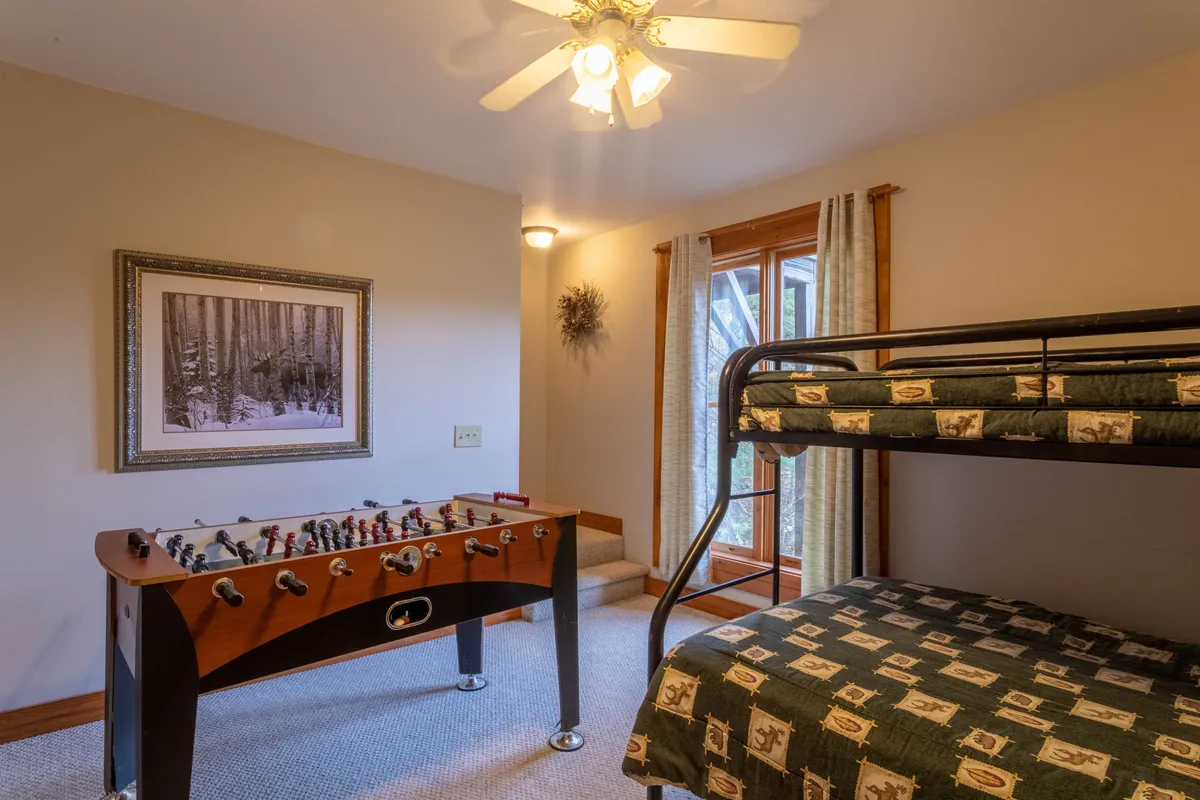 The image showcases a vibrant game room featuring a foosball table as the centerpiece, a sturdy wooden bunk bed in the corner, framed artwork on the walls, and large windows that let in natural light. The space feels lively and playful, inviting guests to engage in games or relax by the window.