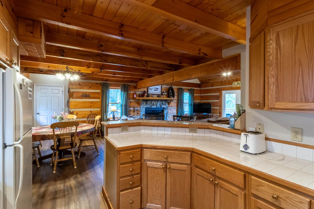 An open-concept view highlighting the kitchen island, wooden beams overhead, a spacious dining table, and the fireplace beyond. The layout shows how the kitchen, dining, and living spaces are seamlessly linked, making it ideal for group meals and celebrations.