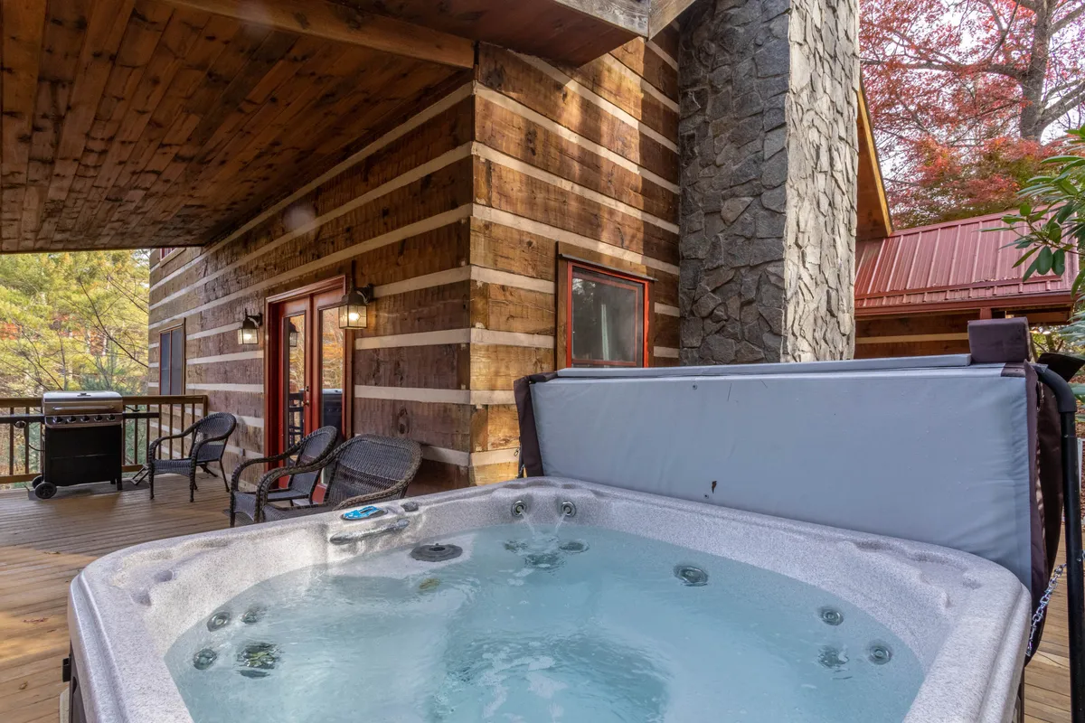 An inviting covered deck featuring a bubbling hot tub, wooden decking, sturdy stone columns, and surrounded by lush trees. The hot tub is positioned to offer stunning views of the forested hills, while the ample deck space suggests room for outdoor dining and relaxation.