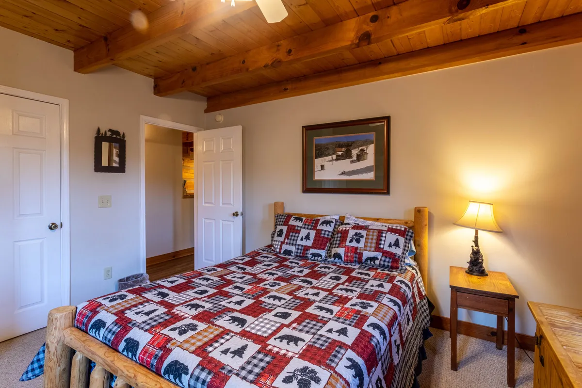 A serene bedroom with a cozy bed dressed in a colorful quilt, wooden ceiling beams, a nightstand with a lamp, and tasteful artwork on the wall. The ambiance is warm and restful, ideal for unwinding.