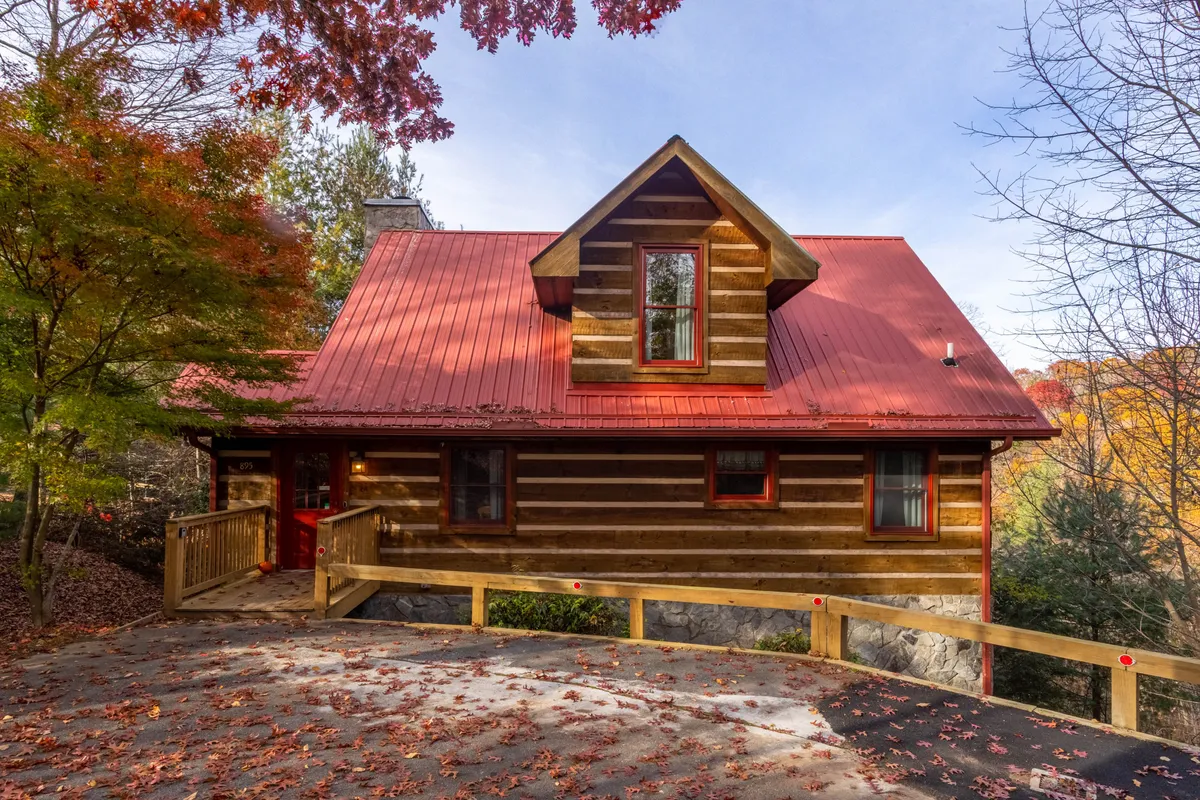 A wide view of the cabin's exterior, showcasing the log structure, red metal roof, wooden railings, and the surrounding trees. The spacious porch hints at outdoor living and offers a sense of connection between the home and its mountain setting.