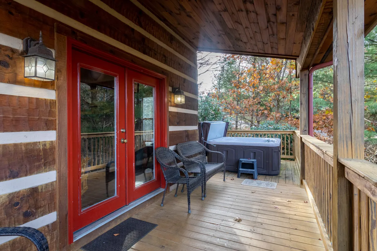 A charming outdoor space with red double doors leading to the deck, a prominently placed hot tub, and woven chairs set up for relaxation. The scene exudes comfort and a cozy vibe, with nature visible in the background, inviting guests to soak or socialize under the sky.