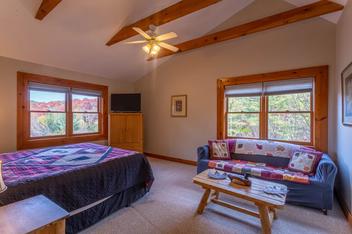 A cozy interior with a comfortable bed topped with a patterned quilt, a sofa adorned with decorative pillows, and large windows that let in plenty of natural light. Wooden furniture and soft lighting create a homely and inviting atmosphere, perfect for relaxing, watching TV, or enjoying views outside.