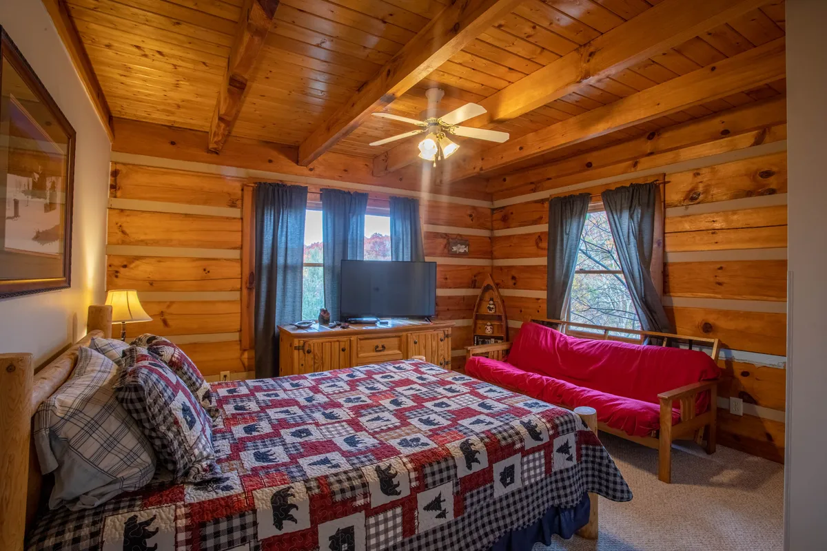 A spacious bedroom featuring a bed with a patterned quilt, inviting wooden furniture, a window with a scenic view, a TV, and a sofa bed. Natural light streams in, highlighting the cozy textures and thoughtful layout ideal for relaxation or entertainment.