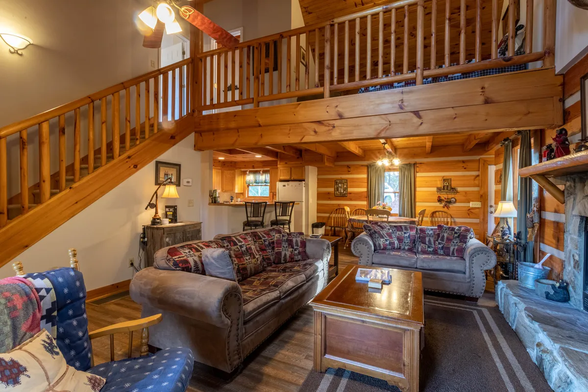 A welcoming living space showcases comfortable sofas, a prominent stone fireplace, a wooden staircase, and a glimpse of the adjacent dining area. The arrangement encourages socializing and relaxation, perfect for movie nights or unwinding after mountain adventures.