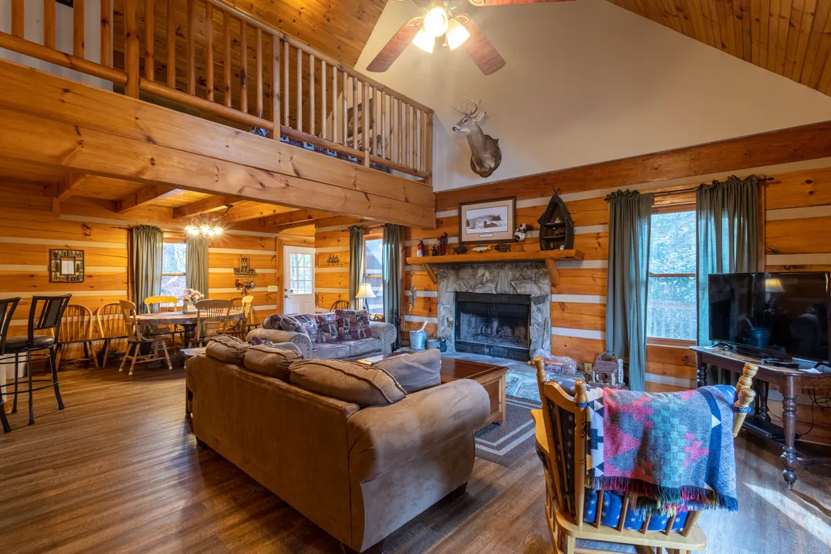This image features the living room with a prominent stone fireplace, cozy sofas, a dining area in the background, lofted ceilings with exposed beams, and inviting decorative touches. The view captures a sense of openness and warmth, aligning with the grand gathering atmosphere described.